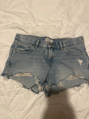 Blå jeansshorts från Perfect Jeans - Snygga blå jeansshorts från Perfect Jeans med råa kanter och slitna detaljer framtill. Klassisk femficksmodell med knapp och dragkedja. Perfekta för varma dagar och en avslappnad stil.
