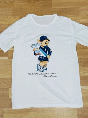 Vit Polo Bear t-shirt från Ralph Lauren - Vit t-shirt från Ralph Lauren med ikoniska Polo Bear-trycket framtill. Björnen har på sig en blå outfit och keps, och under motivet står det 'POLO BEAR by RALPH LAUREN'. Perfekt för en avslappnad och stilren look.