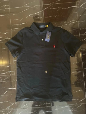 Svart pikéskjorta Polo Ralph Lauren - Svart pikéskjorta från Polo Ralph Lauren i modellen Custom Slim Fit. Skjortan har klassisk krage, två knappar och den ikoniska röda broderade loggan på bröstet. Tillverkad i bomull för en bekväm och stilren känsla.