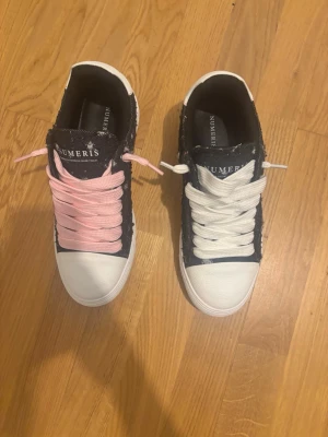 Numeris sneakers med olika snören - Snygga sneakers från Numeris med svart ovandel, vit tå och sula. Ena skon har rosa snören och den andra vita, vilket ger en unik och trendig look. Skorna är låga och har en klassisk rund tå. Materialet ser ut att vara en mix av textil och syntet.