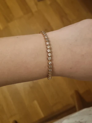 Guldigt tennisarmband med stenar - Snyggt tennisarmband i guldton med runda, klara stenar längs hela bandet. Armbandet har en smal och elegant design med diskret lås. Perfekt för dig som gillar stilrena och glittriga accessoarer.