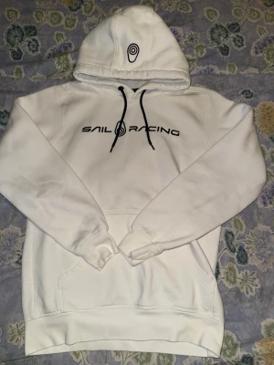 Vit hoodie från Sail Racing - Vit hoodie från Sail Racing med svart logga och text på bröstet. Klassisk känguruficka framtill, svarta dragsnören i huvan och tryckt emblem på huvan. Mjuk och skön bomullsblandning, perfekt för en avslappnad stil.