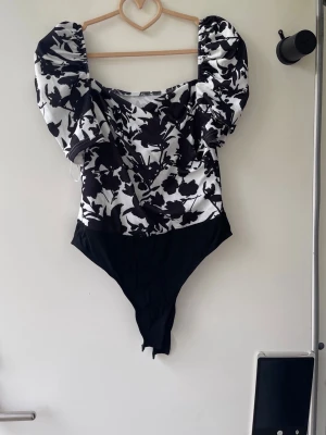 Svartvit body med puffärmar - Trendig body med svartvitt blommönster och stora puffärmar. Djup, fyrkantig urringning och enfärgad svart nedre del. Perfekt för dig som vill sticka ut med en cool och modern look. Materialet ser ut att vara stretchigt och bekvämt.helt ny
