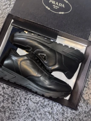 Svarta Prada sneakers - Snygga svarta sneakers från Prada i äkta läder med robust sula och diskret logga på sidan. Skorna har en klassisk låg profil och stilrena detaljer som passar perfekt till streetwear eller en mer dressad look.
