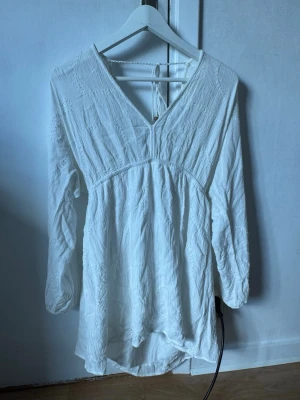 Vit klänning med v-ringning - Vit kortklänning med v-ringning och långa ärmar. Klänningen har broderade detaljer och knytning i nacken. Tillverkad i ett lätt bomullstyg med struktur, perfekt för varmare dagar.