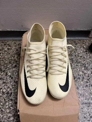 Nike Mercurial vita fotbollsskor - Snygga vita Nike Mercurial fotbollsskor med svart swoosh-logga på sidan och texten 'Mercurial' bak. Skorna har högt skaft och snörning framtill. Tillverkade i syntetmaterial för bra grepp och känsla på planen.