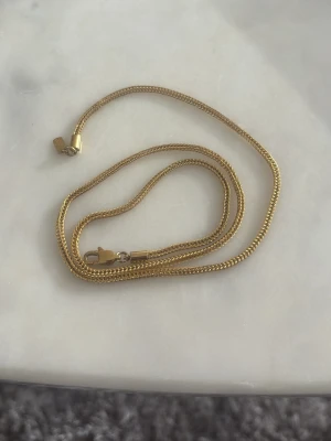Guldfärgat kedjehalsband - Snyggt guldfärgat halsband med klassisk kedjedesign. Halsbandet har en smal, platt kedja och ett stilrent lås i metall. Perfekt för att ge en elegant touch till din outfit. Passar både till lager-på-lager och som ensam accessoar.