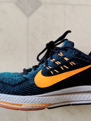 Nike Zoom Structure 18 Man - Snygga sneakers från Nike i blått och svart med orange detaljer och stor orange swoosh-logga. Ovandelen är i mesh för bra andningsförmåga och sulan har Zoom-dämpning för extra komfort. Perfekta för dig som gillar sportig stil och vill sticka ut. Skorna är i bra skick. Storlek 42.5