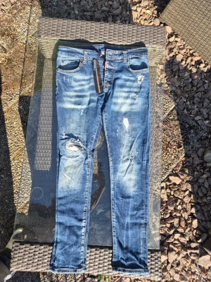 Blå sjeans från Dsquared2 - Säljer ett par blå jeans från Dsquared2 med slitningar och hål på benen. Jeansen har skinny fit, dragkedjegylf och klassiska fem fickor. Snygga detaljer med ljusa partier och råa kanter. Perfekta för en trendig streetstil.
