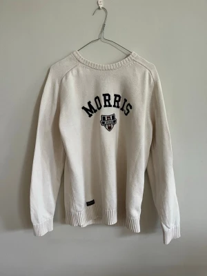 Vit tröja från Morris  - Vit långärmad tröja från Morris med mörkblå broderad logga och emblem på bröstet. Tröjan har ribbade muddar vid ärmslut och nederkant samt rund halsringning. Perfekt för en stilren och sportig look.