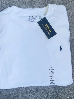 Vit t-shirt från Polo Ralph Lauren - Klassisk vit t-shirt från Polo Ralph Lauren med rund hals och korta ärmar. T-shirten har den ikoniska blå logotypen broderad på bröstet och är tillverkad i mjuk bomull för en skön känsla. Perfekt basplagg med stilren design.