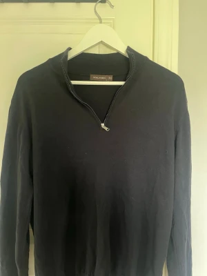 Svart halvzip-tröja från Oscar Jacobson - Stilren svart tröja från Oscar Jacobson med halv dragkedja och ribbad krage. Tillverkad i Italien och passar perfekt för en snygg och avslappnad look. Långärmad modell med klassisk passform.