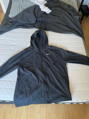 Svart Nike vindbreaker - Säljer en svart hoodie från Nike med dragkedja framtill och huva. Hoodien har två sidofickor och en liten vit Nike-logga på bröstet. Tillverkad i ett lätt och mjukt material som passar perfekt till en avslappnad stil.