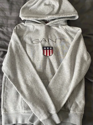 Grå hoodie från GANT med logga - Snygg grå hoodie från GANT med broderad logga och amerikanskinspirerat emblem på bröstet. Hoodien har huva och känguruficka framtill. Tillverkad i mjukt bomullsmaterial, perfekt för en avslappnad och sportig stil.