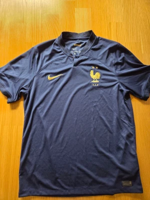 Nike Frankrike dam fotbollströja, storlek L - Mörkblå fotbollströja med korta ärmar och en krage med en knappstängning. Tröjan har en guldfärgad Nike-logotyp på höger bröst och Frankrikes fotbollsförbunds emblem med två stjärnor och texten "FFF" på vänster bröst. Ärmarna har detaljerade mönster och en liten rand i rött, vitt och blått på ena ärmkanten. Insidan av kragen har texten "NOS DIFFÉRENCES NOUS UNISSENT" i guld. Från säsong 2022-2023 Helt ny dock utan papperslappar
