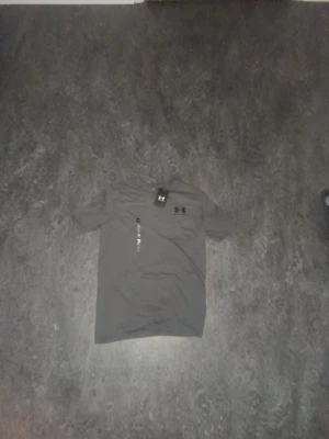 Grå t-shirt från Under Armour - Snygg grå t-shirt från Under Armour med klassisk logga på bröstet. T-shirten har rund halsringning och korta ärmar, perfekt för en sportig och avslappnad stil. Materialet känns mjukt och lätt mot huden.