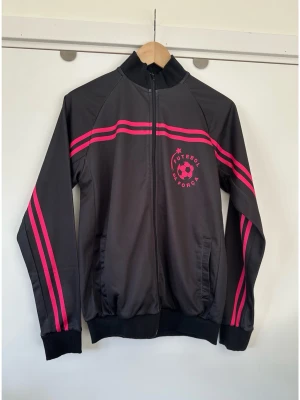Svart träningsjacka med rosa detaljer, storlek 38 - Svart träningsjacka från Futebol da Forca med rosa ränder längs ärmar och bröst, samt tryck på bröstet och ryggen. Jackan har dragkedja, hög krage och muddar vid ärmslut och midja. Perfekt för fotboll eller träning.