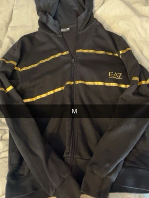 Svart EA7 hoodie med gula ränder - Svart hoodie från EA7 med dragkedja och huva. Snygga gula ränder över bröstet och armarna samt EA7-logga på vänster sida. Perfekt för en sportig och avslappnad stil.