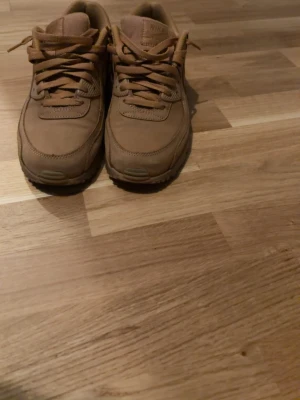 Nike Air Max 90 bruna beige sneakers - Snygga Nike Air Max 90 sneakers i helbrunt läder och mocka. Klassisk siluett med synlig Air Max-dämpning i sulan och diskret Nike-logga på sidan. Skorna har matchande bruna skosnören och en robust yttersula för extra komfort.
