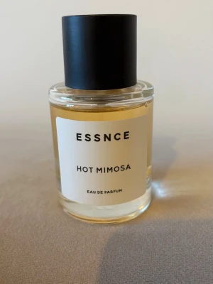 ESSNCE Hot Mimosa Eau de Parfum - ESSNCE Hot Mimosa Eau de Parfum i en stilren, rund glasflaska med svart lock. Parfymen har en ljusgul nyans och etiketten är enkel med svart text. Perfekt för dig som gillar fräscha och blommiga dofter med en modern touch.