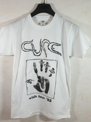 Vit The Cure Wish Tour '92 t-shirt - Säljer en vit t-shirt från Fruit of the Loom med tryck från The Cure Wish Tour '92. T-shirten har svart grafiskt tryck med bandets logga och en hand med ögon. Klassisk rund hals och korta ärmar. Perfekt för dig som gillar bandtröjor. Storlek: Smal