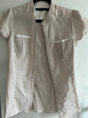 Beige kortärmad blus från Zara Basic - Stilren beige blus från Zara Basic med diskreta smala ränder, korta puffärmar och två bröstfickor med satinliknande kant. Blusen har ståkrage och knäppning framtill. Perfekt för en trendig och avslappnad look.