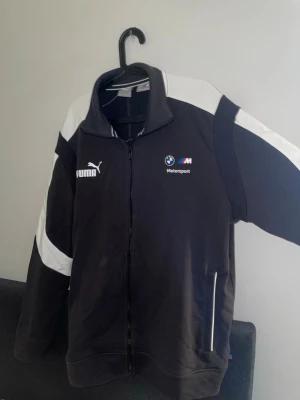 Svart BMW Puma ziptröja - Svart ziptröja från Puma i samarbete med BMW Motorsport. Tröjan har vita detaljer på axlar och ärmar, hög krage och dragkedja framtill. Broderade loggor från både Puma och BMW M på bröstet. Perfekt för dig som gillar sportig stil. Storlek XL