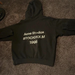 Snygg acne hoodie - Säljer en svart hoodie med trycket 'Acne Studios STOCKHOLM 1996' på ryggen. Hoodien har en klassisk huva och ribbade muddar vid ärmslut och nederkant. Perfekt för dig som gillar streetwear och enkel design.