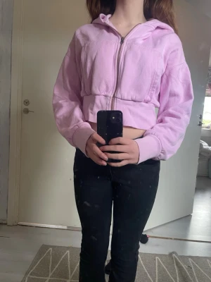 Ljuslila cropped hoodie med dragkedja - Säljer en ljuslila cropped hoodie med dragkedja framtill och huva. Hoodien har långa ärmar och breda muddar vid ärmslut och nederkant. Perfekt för en chill och avslappnad stil. Materialet känns mjukt och bekvämt mot huden.
