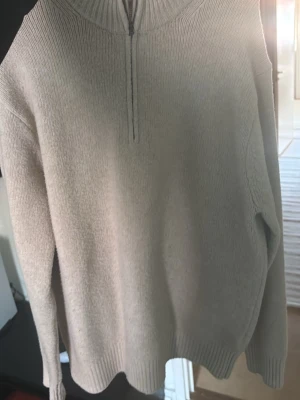 Beige stickad tröja med halv dragkedja från UNIQLO - Säljer en beige stickad tröja med halv dragkedja framtill och ribbade muddar vid ärmslut och nederkant. Tröjan har en hög krage och är perfekt för lager på lager. Enkel och stilren design som passar till det mesta.