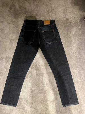 Mörkblå jeans från Nudie Jeans w30 L32 - Säljer ett par mörkblå jeans från Nudie Jeans med klassisk rak passform. Jeansen har kontrastsömmar, två bakfickor och en läderpatch med orange logga bak i midjan. Tillverkade i kraftig denim, perfekta för en stilren vardagslook.