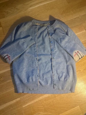 Ljusblå kofta från Burberry - Ljusblå kofta från Burberry med klassiskt rutigt mönster på insidan av knappslån och vid armbågarna. Koftan har ribbade muddar och knäppning framtill. Perfekt för dig som gillar stilrena och exklusiva plagg. Tvättlappens finns ej