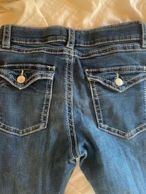 Mörkblå bootcut jeans med ficklock - Snygga mörkblå bootcut jeans med kontrastsömmar och ficklock bak med knappdetaljer. Klassisk femficksmodell och låg midja. Perfekta till en avslappnad stil. Materialet är denim i bomull.