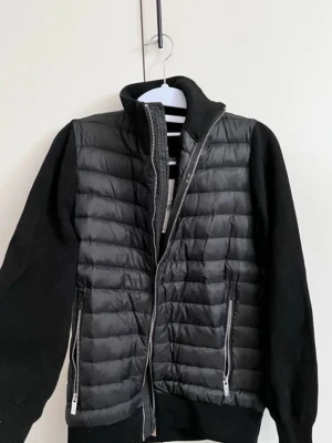 Cardigan från icon Amsterdam  - Använd 1 enstaka gång så i princip helt ny. Stilren svart dunväst med quiltad front och stickade ärmar. Västen har hög krage, två dragkedjefickor och hel dragkedja framtill. Perfekt för lager-på-lager och en modern look. Materialmix av dun och stickat ger både värme och stil.