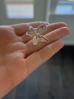 Pilgrim silvrig ring med blomform  - Säljer en snygg silvrig ring från Pilgrim med en stor, detaljrik blomformad dekoration. Ringen har en blank yta och blommans blad är formade i en modern och stilren design. Perfekt accessoar för att lyfta din outfit.