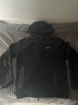 Svart  NOCTA hoodie med dragkedja - Säljer en svart hoodie från  NOCTA med gul logga på bröstet och NOCTA-tryck på ärmen. Hoodien har dragkedja framtill, två fickor med dragkedja och en stor huva. Perfekt för dig som gillar streetwear och stilrena detaljer.