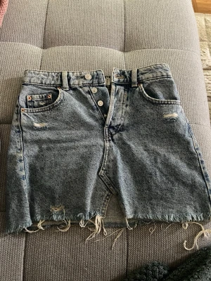 Blå jeanskjol med rå kant från bershka - Trendig blå jeanskjol från Levi's med slitna detaljer och rå, fransig nederkant. Kjolen har knappar framtill och en kort passform. Perfekt för dig som gillar en avslappnad och cool stil. Storlek 32 men skulle säg att den passar 34 också