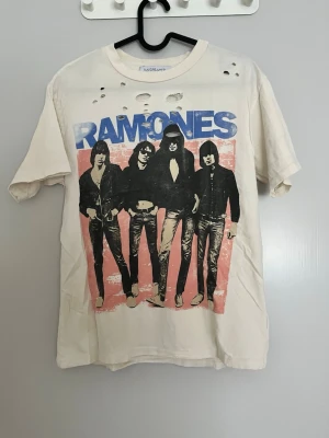 Ramones t-shirt med hål och tryck - Cool vit t-shirt från Rage Age med Ramones-tryck i blått och svart på framsidan. T-shirten har flera hål upptill för en edgy look och klassisk rund hals. Materialet är mjuk bomull och passformen är normal.