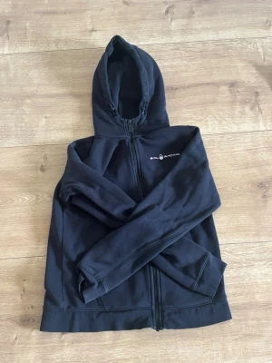 Svart hoodie från Sail Racing - Svart hoodie från Sail Racing med dragkedja framtill och broderad logga på bröstet. Mjuk insida och två fickor. Klassisk modell med huva och ribbade muddar. Perfekt för dig som gillar stilrena och sportiga plagg.