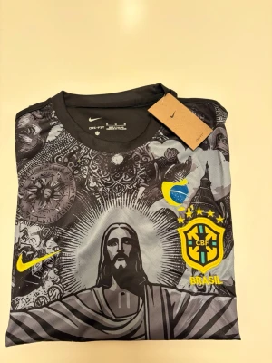 Nike Brasil grafisk fotbollströja - Snygg svart och grå fotbollströja från Nike med Brasil-logga och gult Nike-märke. Tröjan har ett stort grafiskt tryck med Kristusstatyn och brasilianska symboler. Tillverkad i lätt och ventilerande material, perfekt för träning eller match.