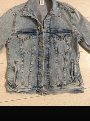 Ljusblå jeansjacka från H&M - Snygg ljusblå jeansjacka från H&M med klassisk krage, bröstfickor med lock och knapp samt sidofickor. Jackan har slitna detaljer och silverfärgade knappar framtill. Perfekt till alla dina outfits för en cool och avslappnad look.