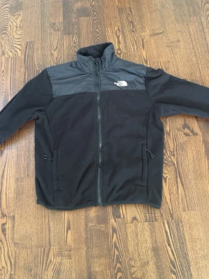 Svart fleecejacka från The North Face - Svart fleecejacka från The North Face med dragkedja framtill och två fickor med dragkedja. Jackan har en hög krage och förstärkt axelparti i nylon. Perfekt för lager-på-lager och outdoor-aktiviteter.