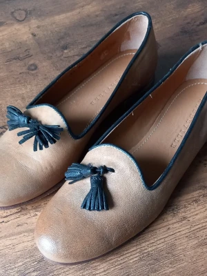 Beige loafers från Alberville med svarta tofsar - Snygga beige loafers från Alberville i läder med svarta tofsar framtill och svarta kanter runt öppningen. Klassisk och stilren design som passar perfekt till jeans eller kostymbyxor.