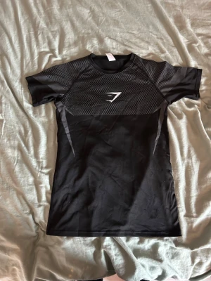 Svart träningströja från Gymshark - Säljer en svart kortärmad träningströja från Gymshark med gråa detaljer och logga på bröstet. Tröjan har meshpaneler upptill för extra ventilation och är tillverkad i ett stretchigt funktionsmaterial som passar perfekt för gym och träning.