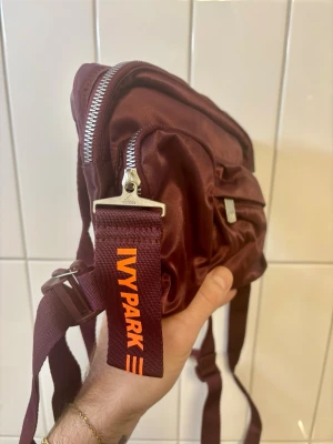 Ivy Park x Adidas x Beyonce  - Snygg vinröd väska/ryggsäck från Ivy Park x Adidas. Väskan är menad att bäras framifrån över bröstet men fungerar även på ryggen. Flera praktiska fack och dragkedjor. Väskan har en cool orange Ivy Park-detalj och är tillverkad i glansigt syntetmaterial. Perfekt för dig som vill ha en trendig och sportig look.                           Aldrig använd! Nypris 2199kr
