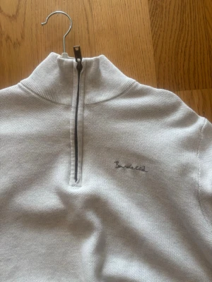 Vit halfzip-tröja  - Vit stickad tröja från  med halv dragkedja och hög krage. Tröjan har diskret broderad logga på bröstet och ribbade muddar vid ärmslut och nederkant. Perfekt för en clean och stilren look.