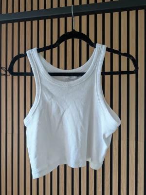 Vit ribbad croppad linnetopp - Snygg vit croppad linnetopp från Bik Bok i ribbat bomullsmaterial. Toppen är ärmlös med breda axelband och har en enkel, stilren design som passar perfekt till jeans eller kjol.