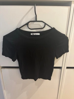 Svart croppad topp från Zara - En svart, croppad topp från Zara med korta ärmar och rund halsringning. Enkel och stilren design som passar perfekt till jeans eller kjol. Tillverkad i mjukt material för en bekväm känsla.