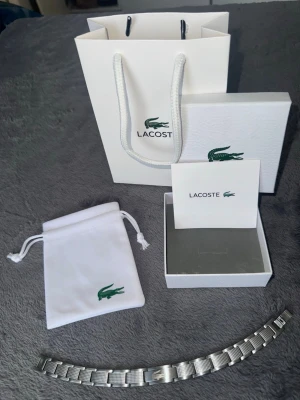Lacoste armband - Stilrent armband från Lacoste i silverfärgat rostfritt stål med klassisk krokodil-logga ingraverad på spännet. Armbandet har en modern, länkad design med diskret rutmönster på sidorna. Perfekt accessoar för dig som gillar minimalistisk och sportig stil.