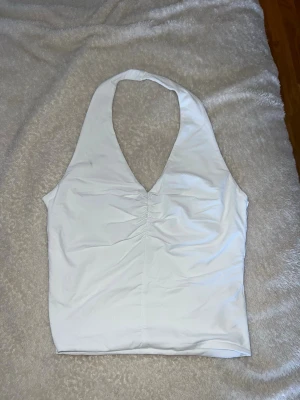 Vit halterneck linnetopp - Säljer en stilren vit halterneck linnetopp med v-ringning och lätt rynkning framtill. Toppen är ärmlös och har en clean, enkel look som passar perfekt till jeans eller kjol. Materialet känns mjukt och stretchigt.